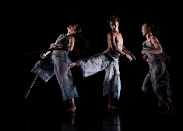3 dancers are on a stage with a black floor and backdrop. 2 female dancers face the same direction. The dancer to the left stands on her leg and holds a crutch in each hand. Her head is tilted backwards and her left crutch is lifted in the air. The other female dancer is running towards her. In between them there is a male dancer facing the opposite direction. He is stood on his left leg and lifts his right leg behind him, touching the dancer behind him. He looks over his shoulder at his lifted foot. The female dancers wear loose- fitting, sheer grey vest tops and trousers and the male dancer wears a similar costume but is shirtless.