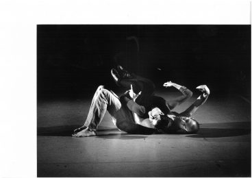 2 dancers lie on the stage floor. One of the dancers legs are bent whilst the other lies on top of them. Both the dancers right arms are lifted in the air.