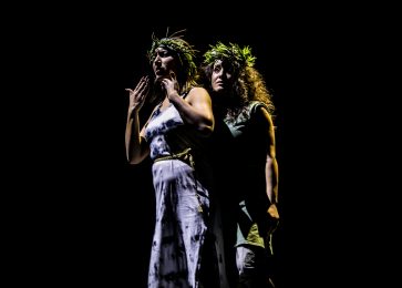 2 people stand next to each other on stage. Oe of them has their hands close to their mouth. They both wear green headpieces.