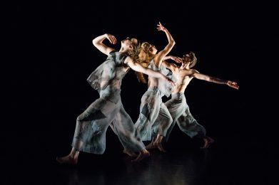 Three dancers in grey transparent clothing, moving past each other, looking up diagonally and arms angled up/outward.