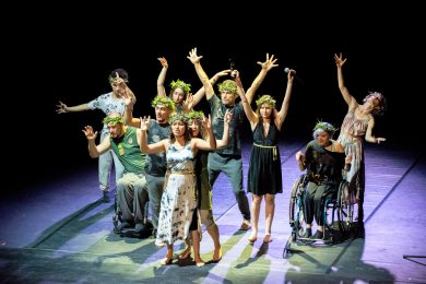 Group of disabled and non-disabled dancers on stage wear leaf headdresses and raise their arms