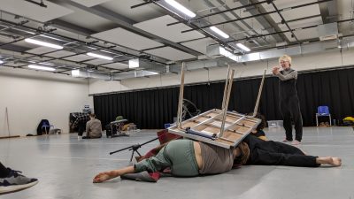 An upside down table is resting on a group of dancers. The back of one dancer can be seen, and their foot is sticking out from under the table.
