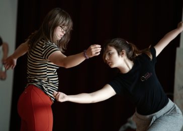Two people dancing in front of one another with their arms stretched sideways.