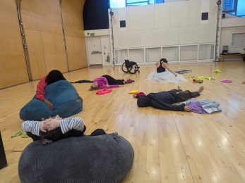 Five dancers resting in a dance studio - some laying and sitting on the wooden floor and some resting on bin bags. Colourful costumes are scattered around the space.