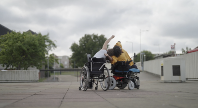 Two people in wheelchairs dancing next to one another outdoors. They lean on each other, one person facing the camera and one facing away from the camera.