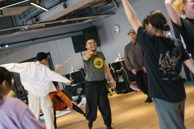 A group of people, in various poses, engage in a dance class. At the centre, Dominic Mitchell leads the class. They raise one arm forward and extend a leg back, smiling joyfully. Others lift their arms or stretch them out, with only two people facing the camera.