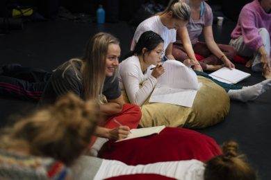 Side view of a group of people leaning on bean bags and taking notes during a workshop. The focus is on one person in the centre, smiling as they write notes and look ahead.