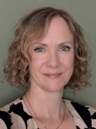 A softly lit headshot of a person with short, blonde curly hair, giving a gentle smile and gazing straight into the camera, with natural tones and a clean pale green background.
