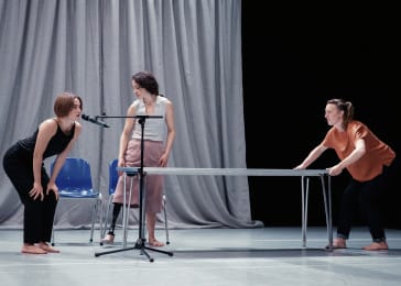 Three performers are on a stage with a grey draped back cloth. There is a table in the middle and one dance is pushing down on the table. Another leans toward a microphone while the last dancer watches on.
