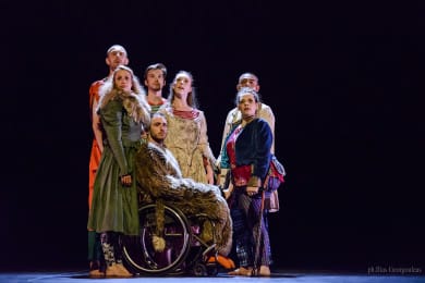 A group of performers in colourful costumes stands closely together on a dark stage, including one performer using a wheelchair, creating a dramatic ensemble portrait.